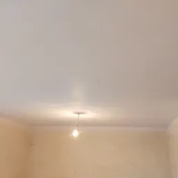 Biały, gładki sufit napinany z widoczną żarówką wiszącą na środku pomieszczenia, ściany przygotowane do malowania.