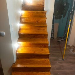 Ihrhause - Odnowione drewniane schody w trakcie prac wykończeniowych, widoczne ślady po remoncie na podłodze i ścianach, w tle drabina i narzędzia.