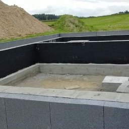 Ihrhause - Budowa fundamentów domu z izolacją przeciwwilgociową, widoczny pracownik w czerwonej kurtce i usypana ziemia w tle.