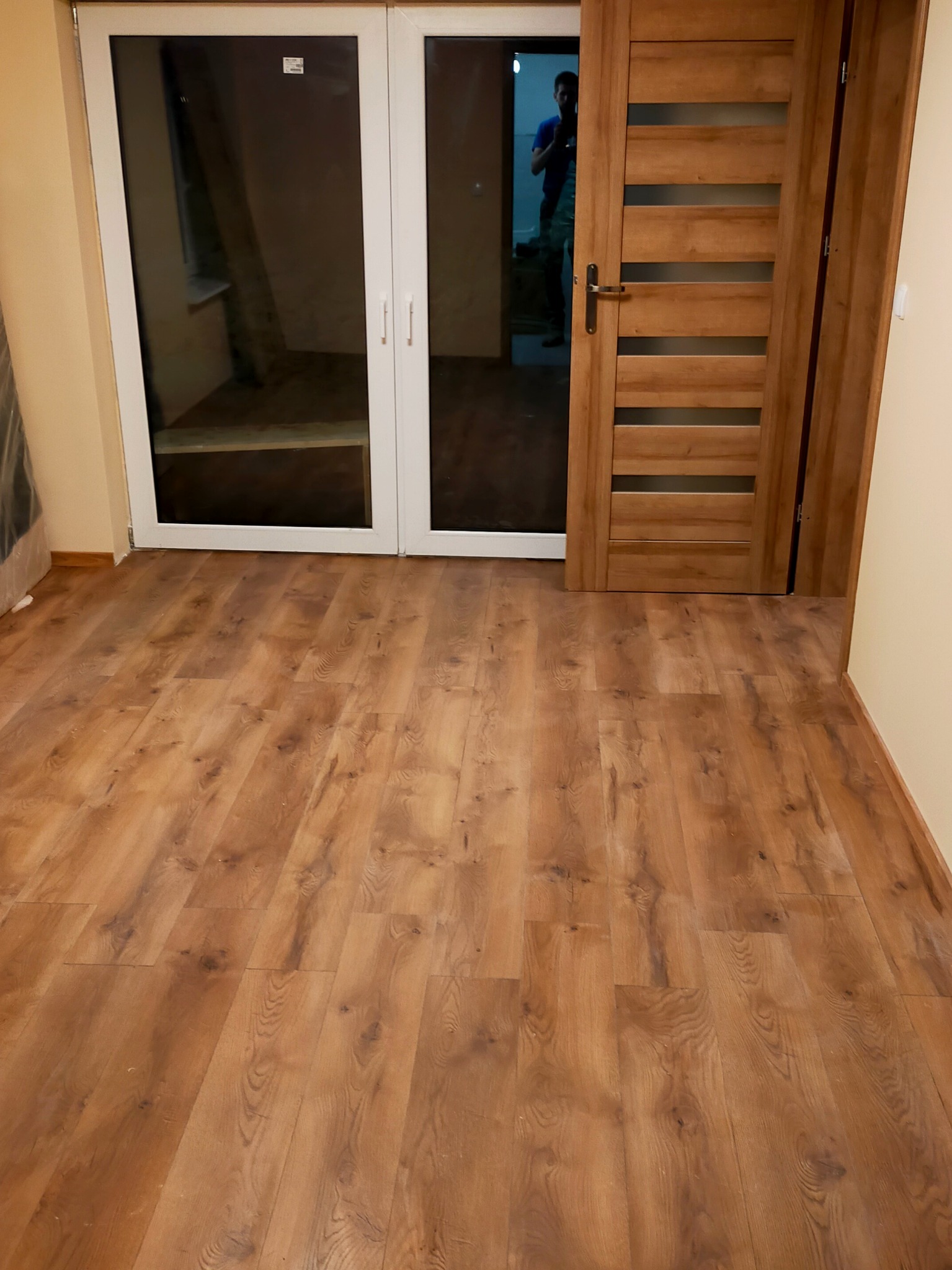 Pokój po remoncie z drewnianą podłogą, białymi drzwiami balkonowymi i drewnianymi drzwiami wewnętrznymi z przeszkleniami, widok na częściowo wyposażone wnętrze.