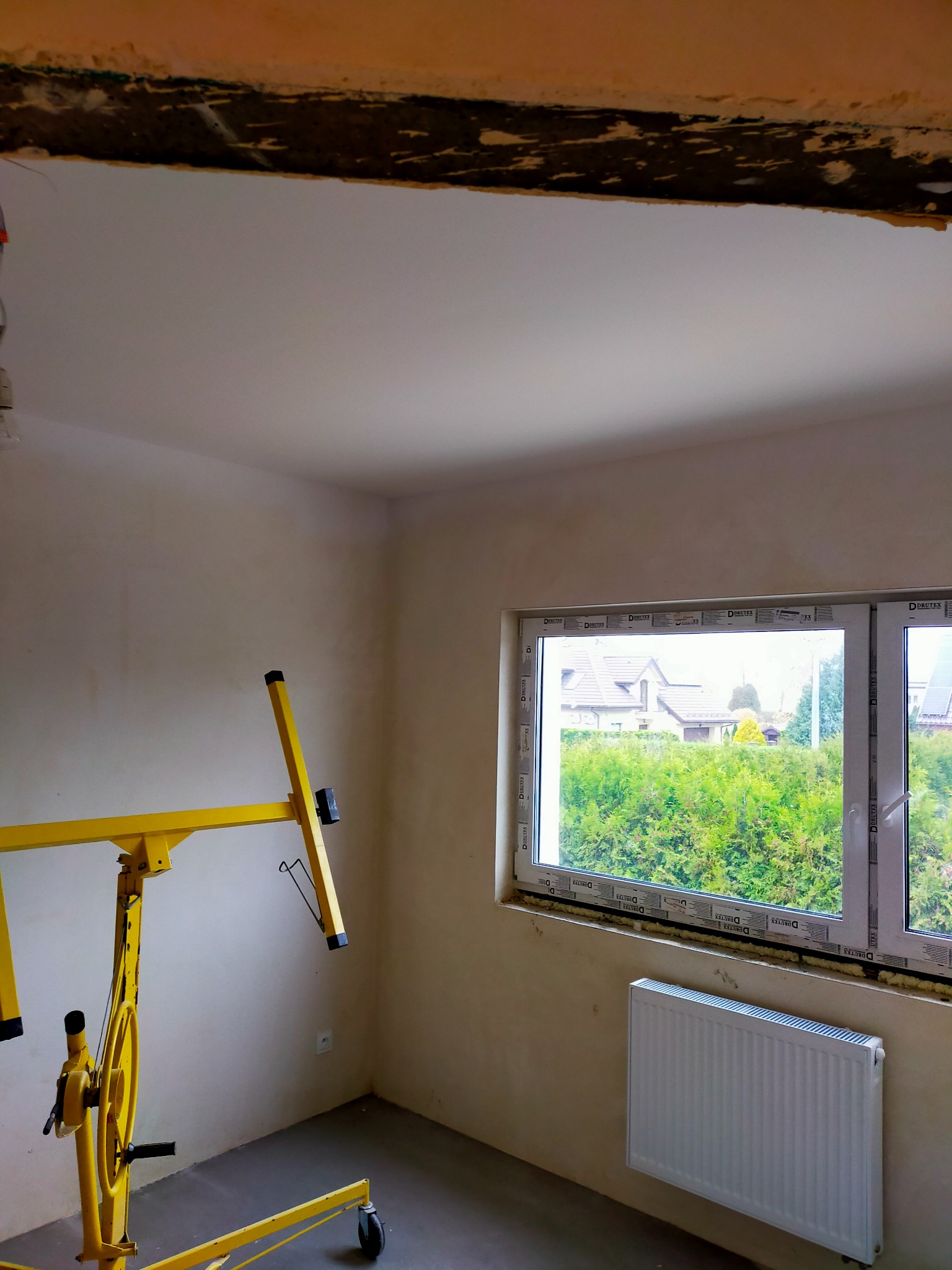 Wnętrze pokoju w trakcie prac wykończeniowych, widoczny podnośnik do płyt gipsowo-kartonowych, okno z widokiem na zieleń i zamontowany grzejnik.