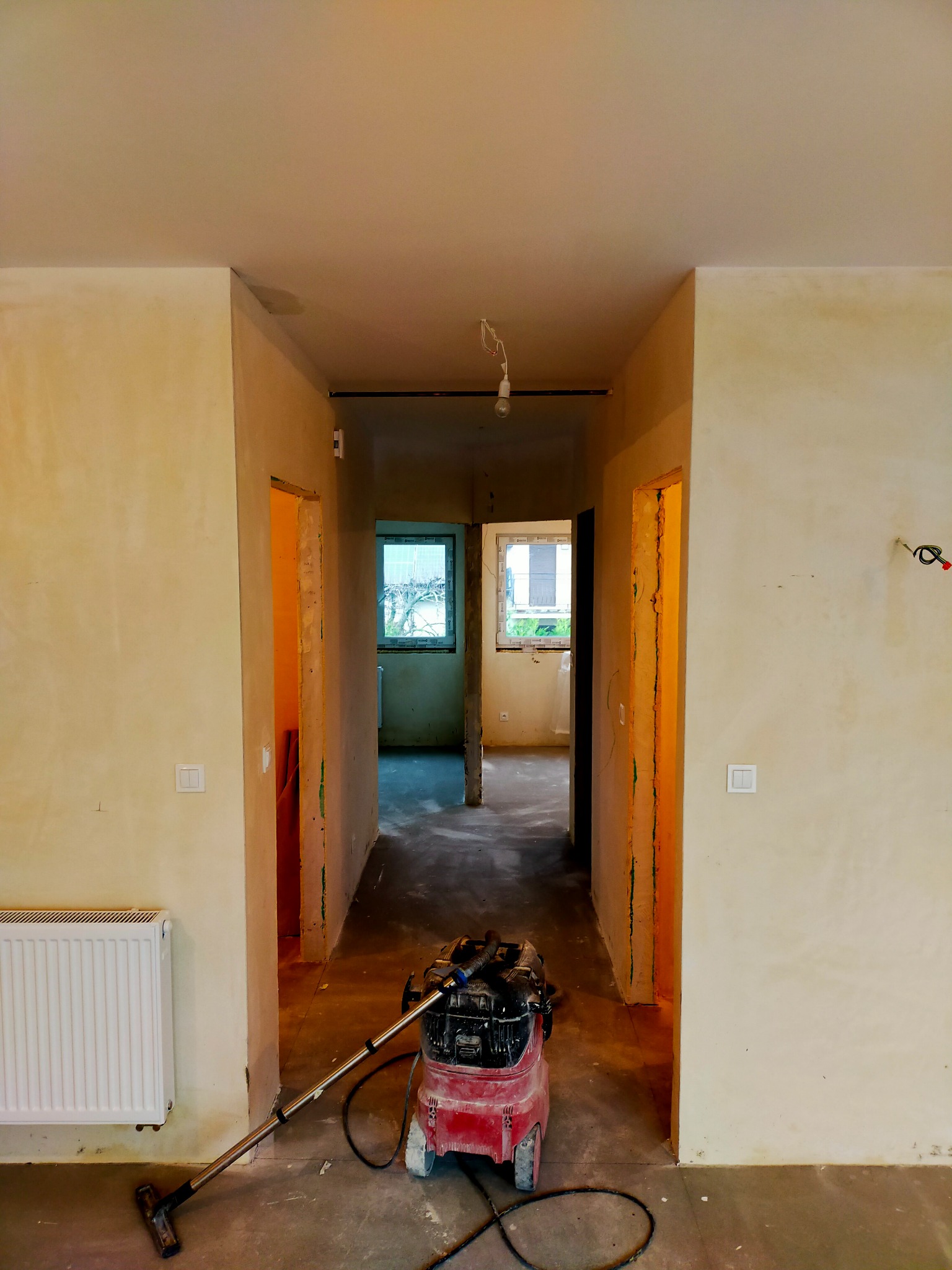 Wnętrze mieszkania w trakcie prac wykończeniowych: widoczne ściany po nałożeniu gładzi, surowy beton na podłodze, grzejnik, odkurzacz przemysłowy oraz przygotowane otwory drzwiowe.
