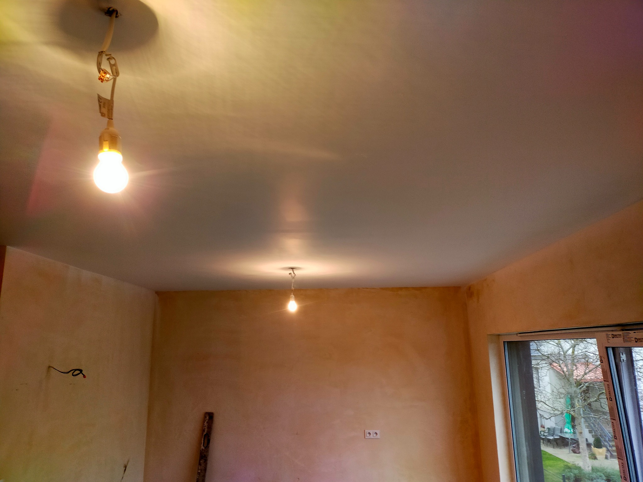 Pokój po nałożeniu gładzi gipsowej, widoczne niedoskonałości na ścianach, sufit z prowizorycznym oświetleniem, okno z widokiem na zewnątrz.