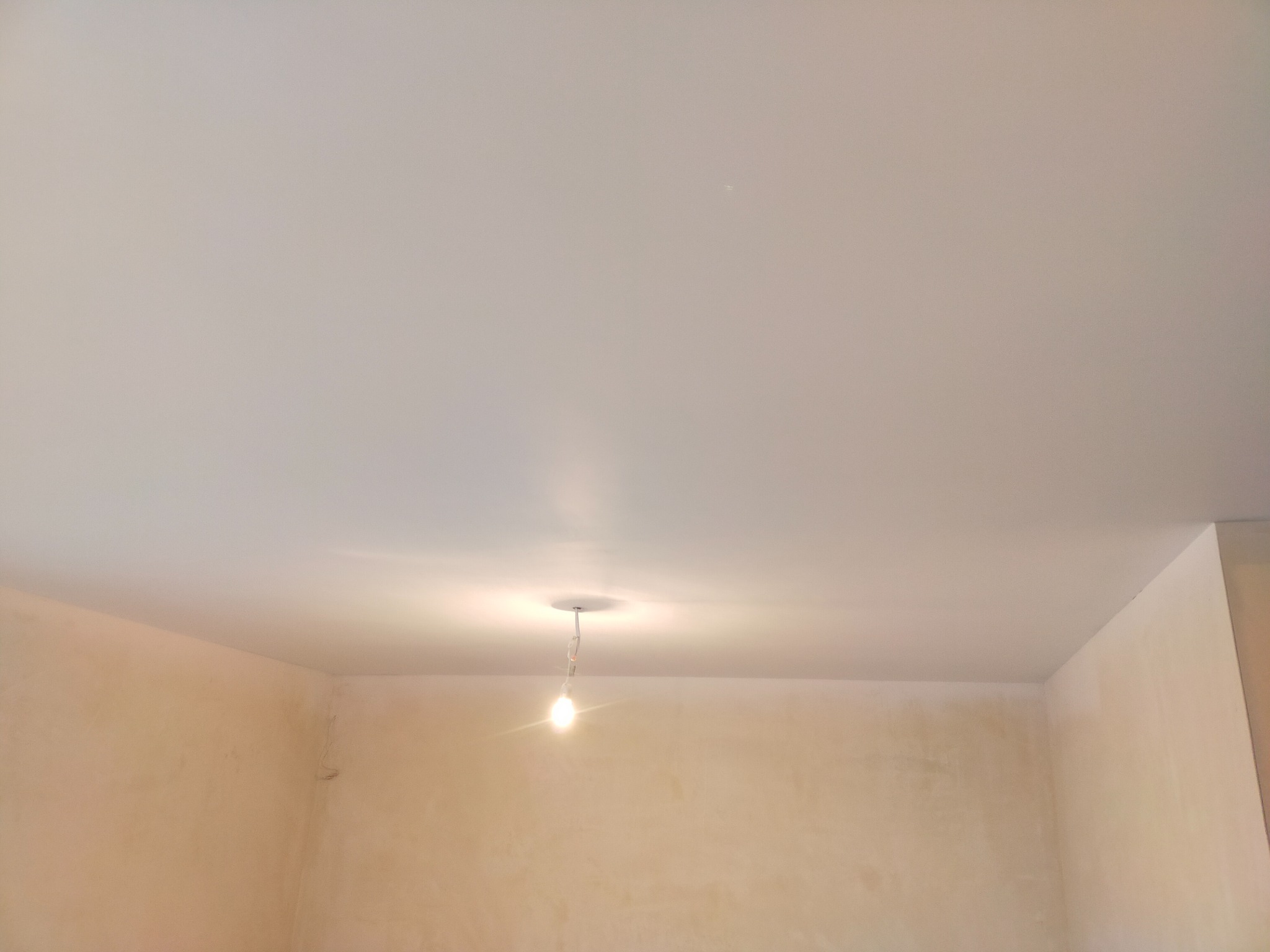 Biały, gładki sufit napinany z widoczną żarówką wiszącą na środku pomieszczenia, ściany przygotowane do malowania.