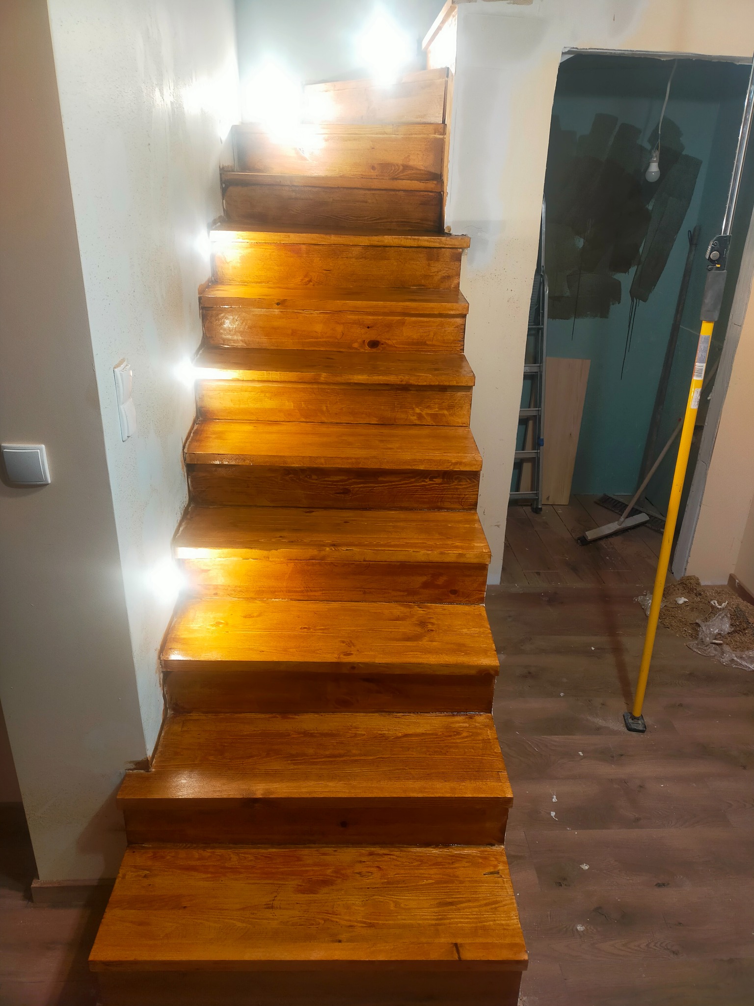 Odnowione drewniane schody w trakcie prac wykończeniowych, widoczne ślady po remoncie na podłodze i ścianach, w tle drabina i narzędzia.