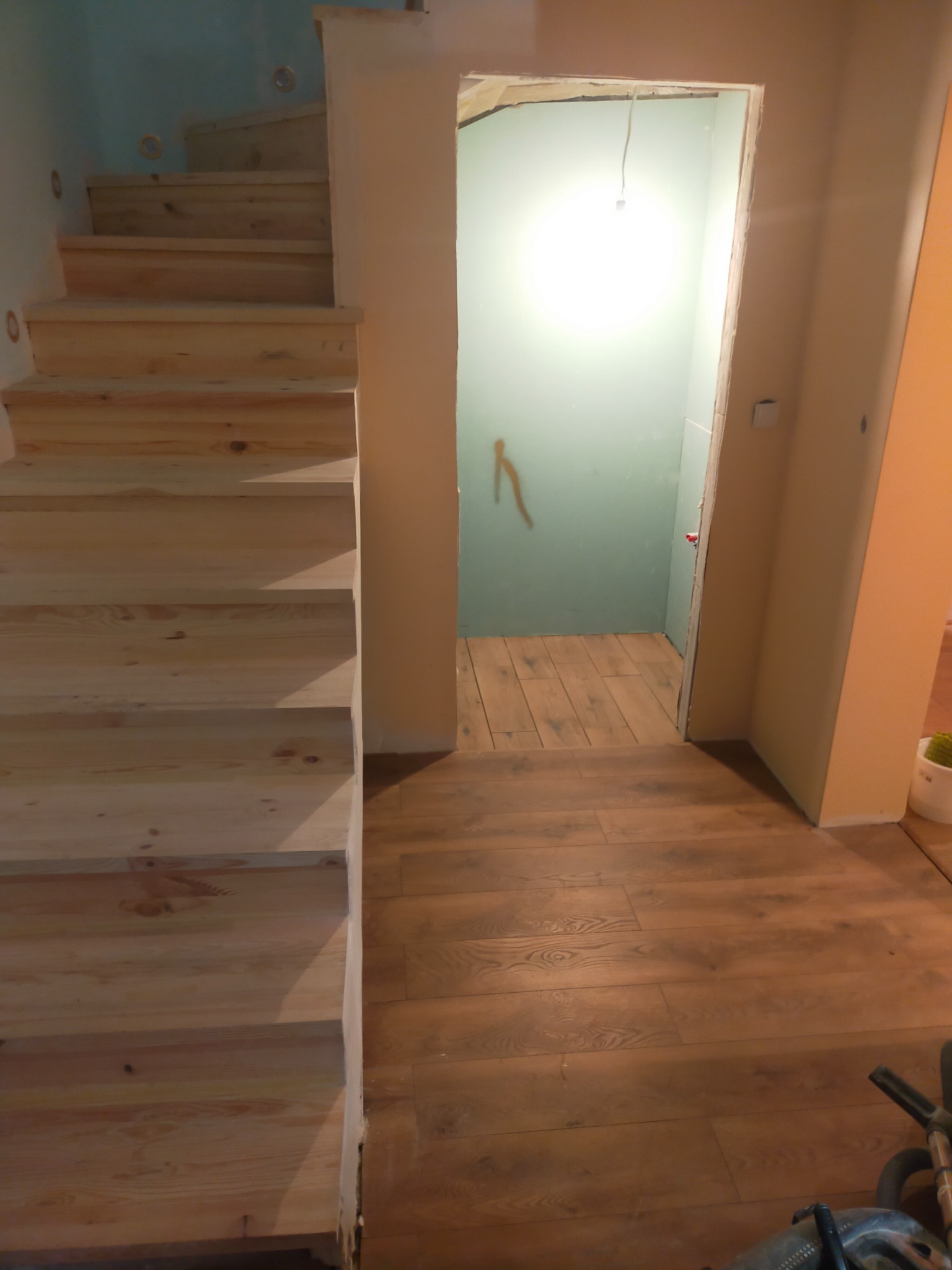 Surowe drewniane schody prowadzą w górę, obok wejście do pomieszczenia z surowymi ścianami i podłogą imitującą drewno, widoczne niedokończone detale.