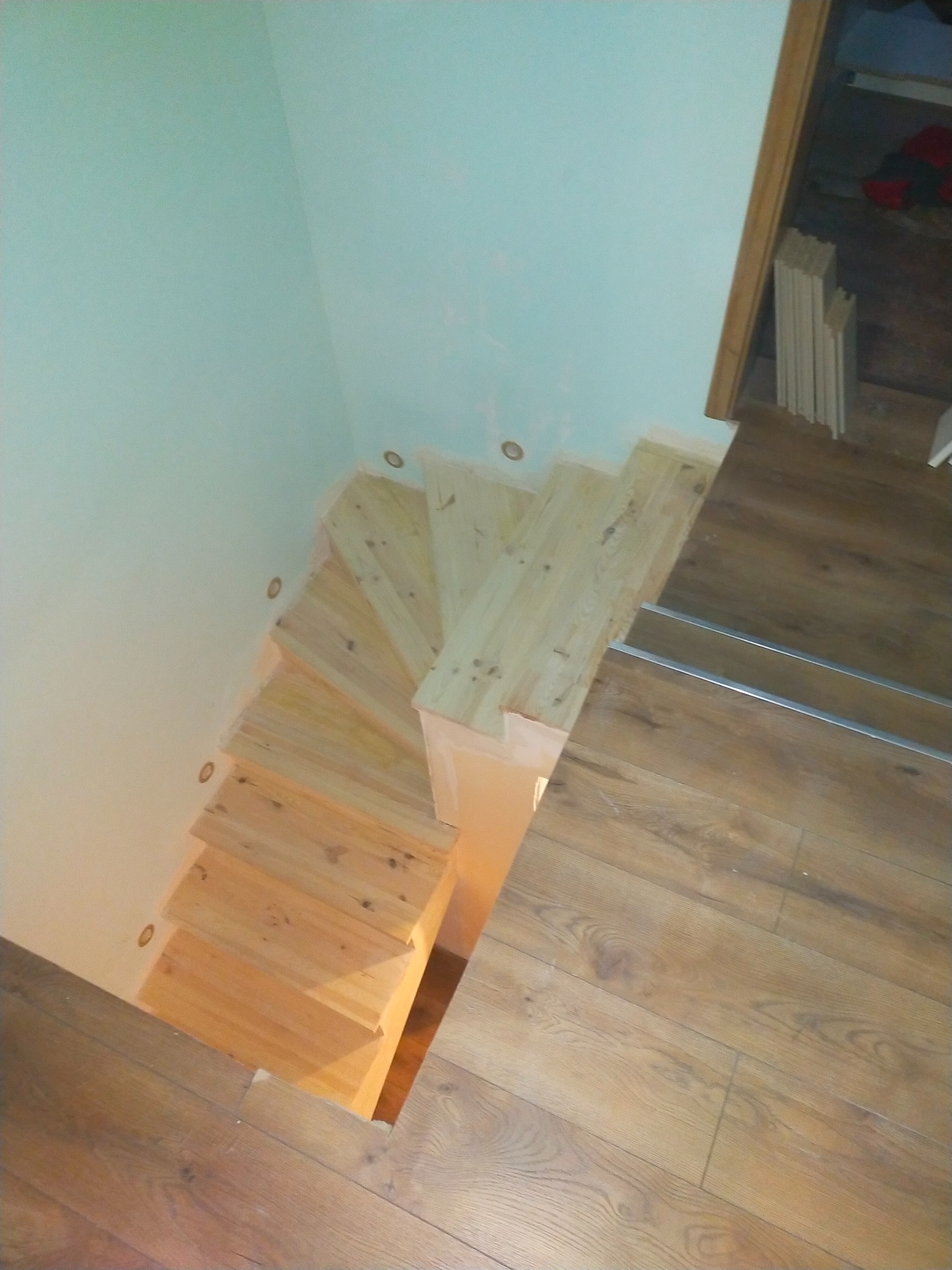 Widok z góry na drewniane schody w trakcie montażu, z widocznymi punktowymi lampkami w ścianie i fragmentem drewnianej podłogi.