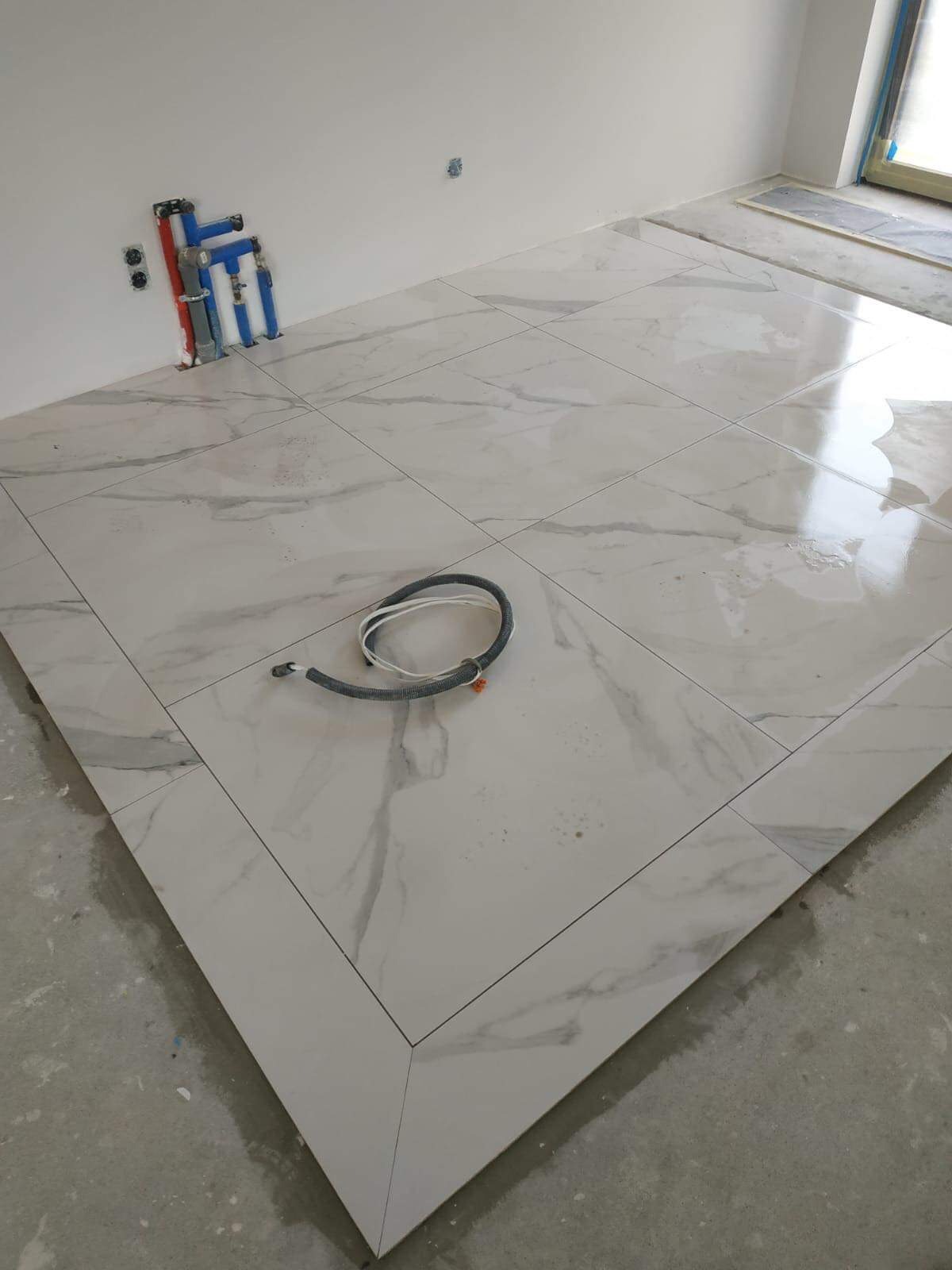 Ułożone płytki podłogowe imitujące marmur z widocznymi instalacjami wodno-kanalizacyjnymi przy ścianie i przewodem elektrycznym na podłodze.