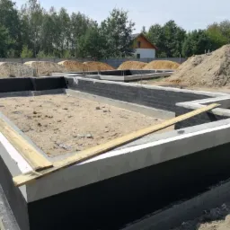 Fundamenty domu w trakcie budowy, z widoczną izolacją przeciwwilgociową i betonową wylewką, na tle hałd piasku i zieleni.