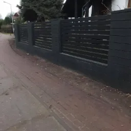 Nowoczesne, ciemnoszare ogrodzenie panelowe z poziomych desek, przylegające do chodnika z czerwonej kostki brukowej, z domem w tle.