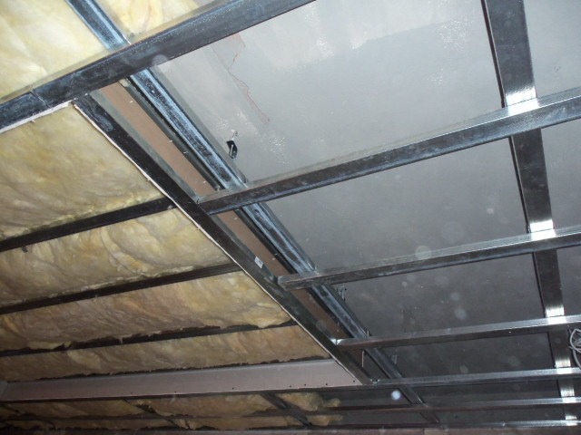 Metalowa konstrukcja sufitu z wełną mineralną i częściowo zamontowaną płytą, widok z dołu.