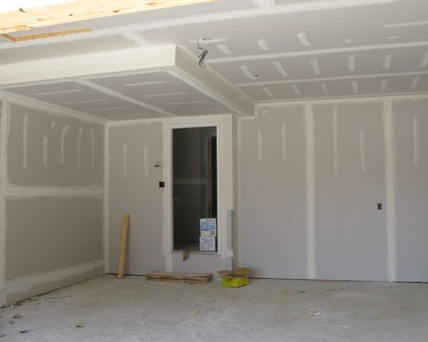 Wnętrze w trakcie wykańczania, ściany i sufit pokryte surowymi płytami gipsowo-kartonowymi ze widocznymi łączeniami, niezabezpieczony otwór drzwiowy, podłoga betonowa, widoczne przewody elektryczne.