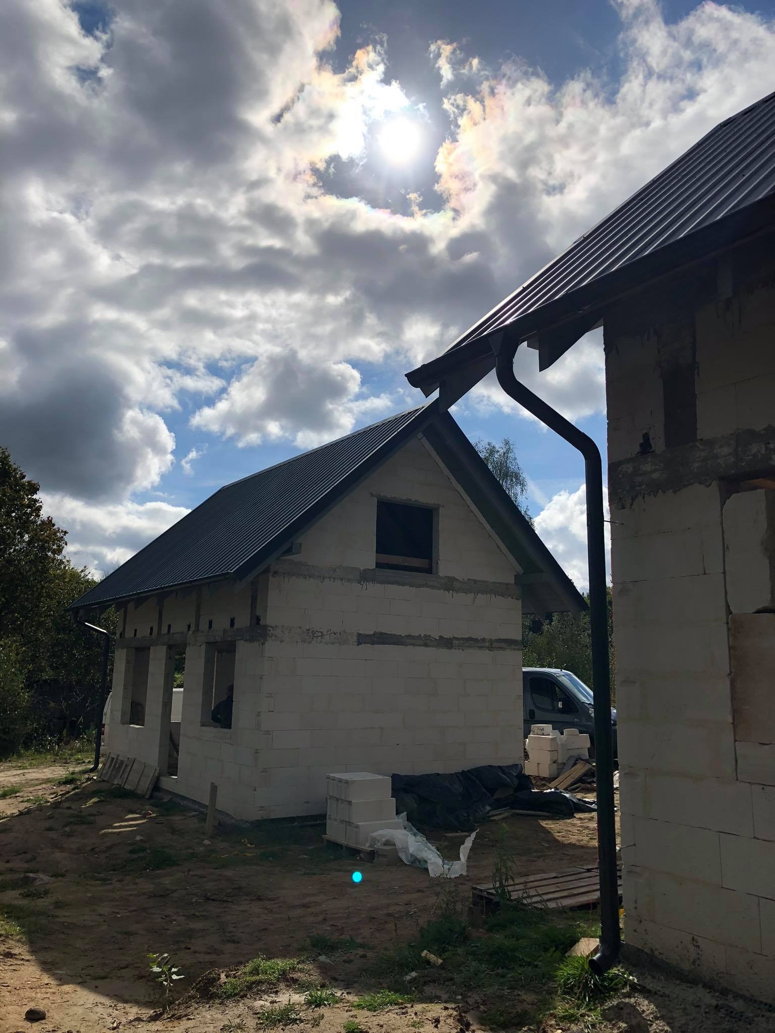 Surowy stan budynku z białych pustaków z ciemnym dachem blachodachówką, widoczne okna bez szyb, w tle błękitne niebo z chmurami i fragment lasu.