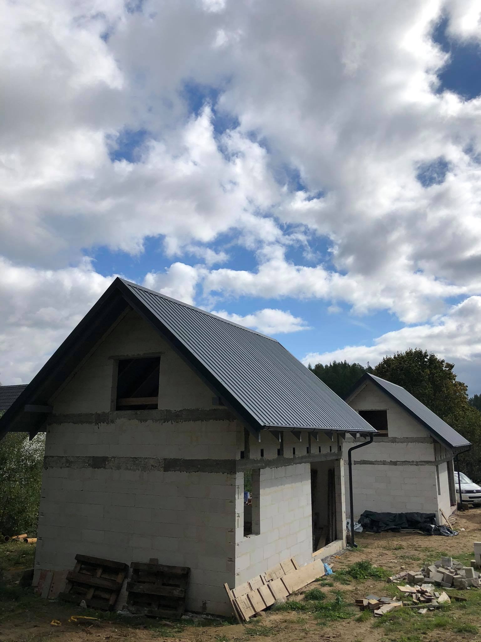 Dwa surowe budynki w stanie deweloperskim z szarym blaszanym dachem na tle błękitnego nieba z chmurami.