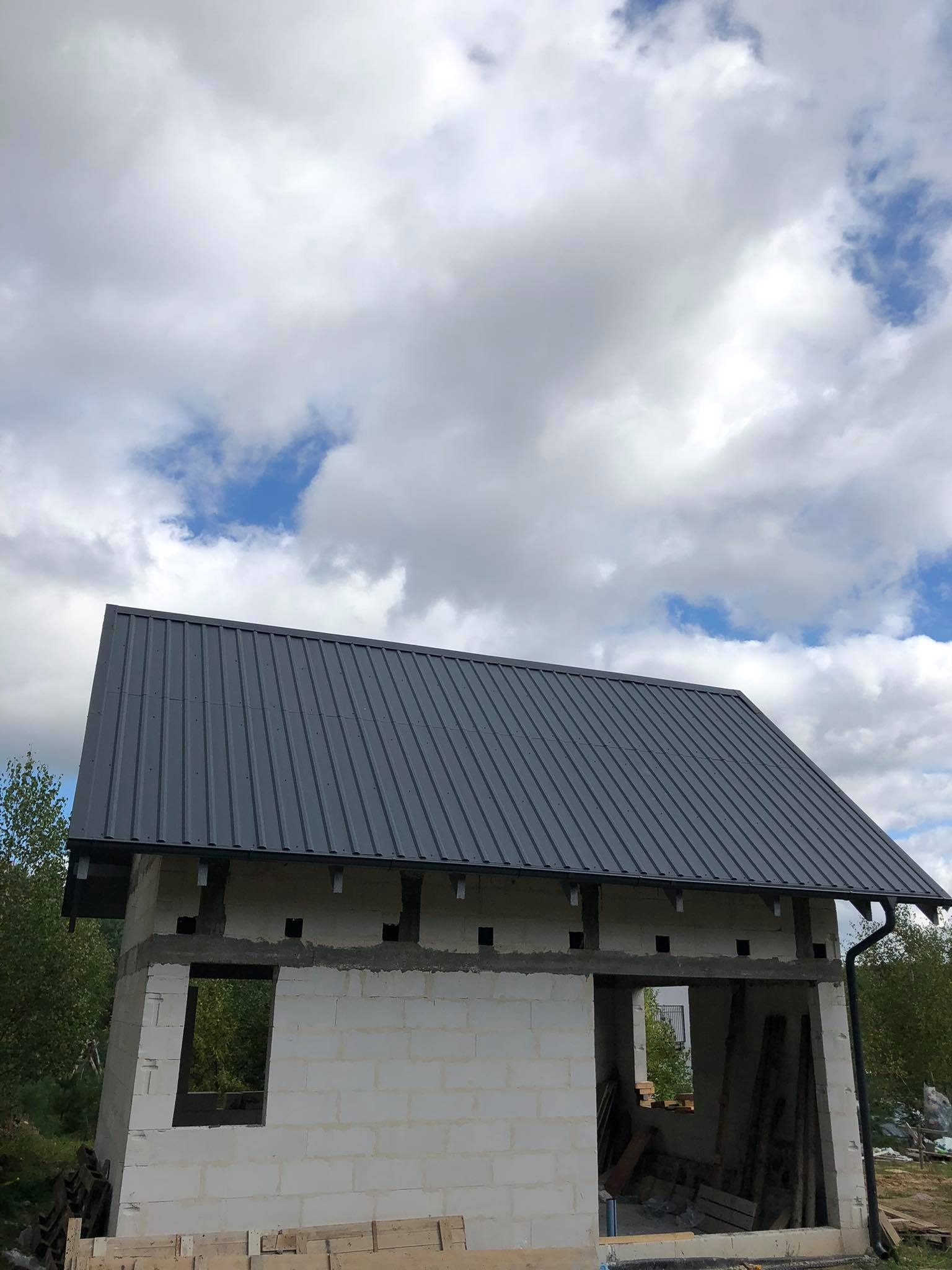 Surowy budynek jednorodzinny z szarej blachy trapezowej na dachu, widoczne otwory okienne i drzwiowe, w tle błękitne niebo z chmurami.