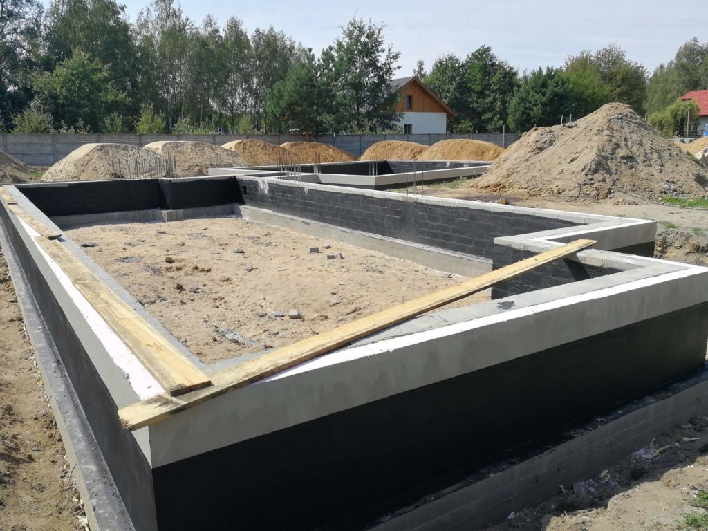 Fundamenty domu w trakcie budowy, z widoczną izolacją przeciwwilgociową i betonową wylewką, na tle hałd piasku i zieleni.