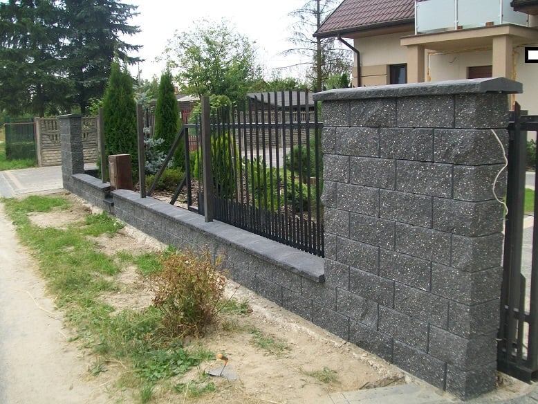 Nowoczesne ogrodzenie z szarego kamienia łupanego z metalowymi przęsłami na tle domu jednorodzinnego z zielonym ogrodem.