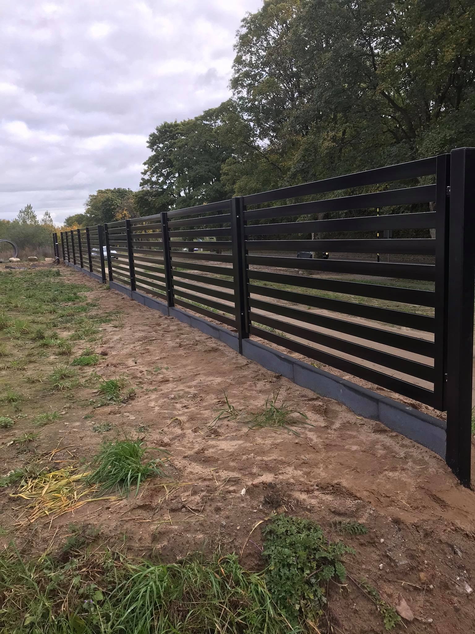 Nowoczesne, poziome, czarne ogrodzenie panelowe z betonową podmurówką na tle zieleni i pochmurnego nieba.