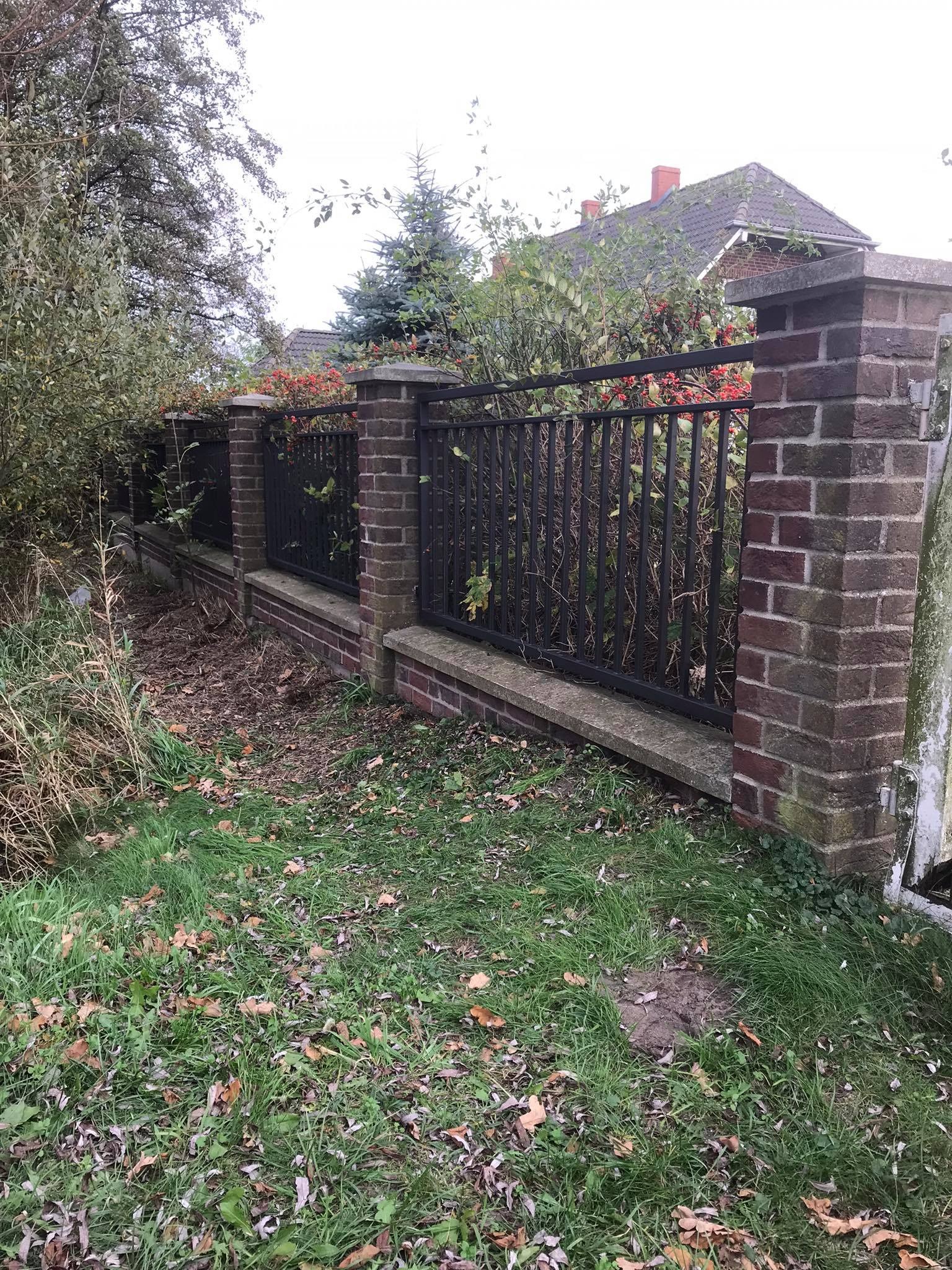 Fragment ogrodzenia z ceglanymi słupkami i metalowymi przęsłami, zarośnięty roślinnością z widocznym budynkiem w tle.