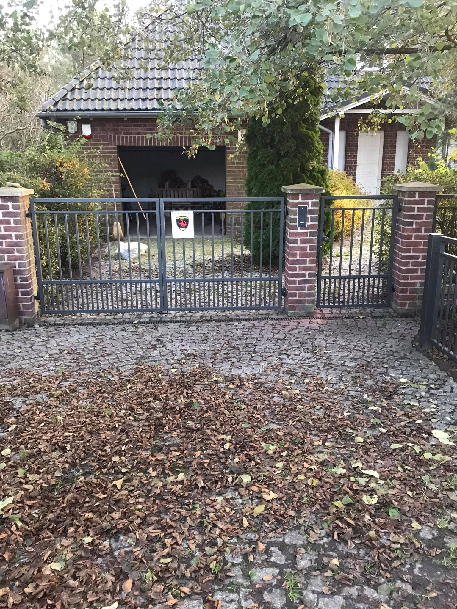 Metalowa brama wjazdowa z ceglanymi słupkami prowadząca do posesji z garażem, na brukowanym podjeździe leżą opadłe liście.