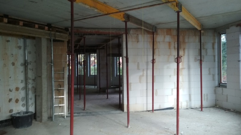 Wnętrze w trakcie budowy z widocznymi podporami stropowymi, pustakami, drabiną i czarnym wiadrem, ślady po zalaniach na ścianach.