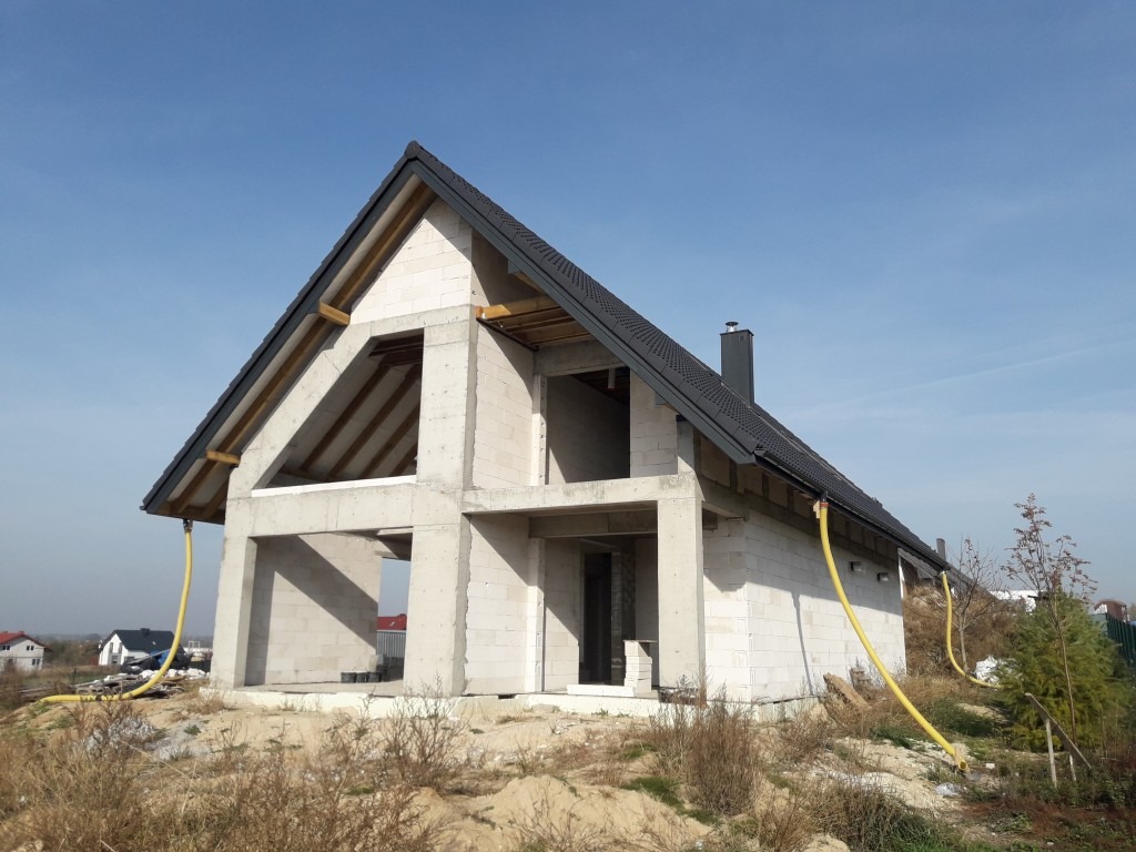 Surowy stan domu jednorodzinnego z betonu komórkowego pod czarnym dachem, widoczne żółte węże na elewacji, w tle zabudowa jednorodzinna.