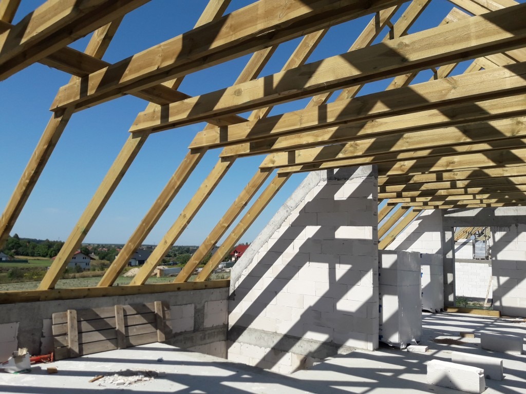 Surowe drewniane krokwie dachowe w konstrukcji domu jednorodzinnego w stanie surowym otwartym, widoczne białe bloczki betonu komórkowego oraz elementy szalunku, w tle błękitne niebo i zabudowa...