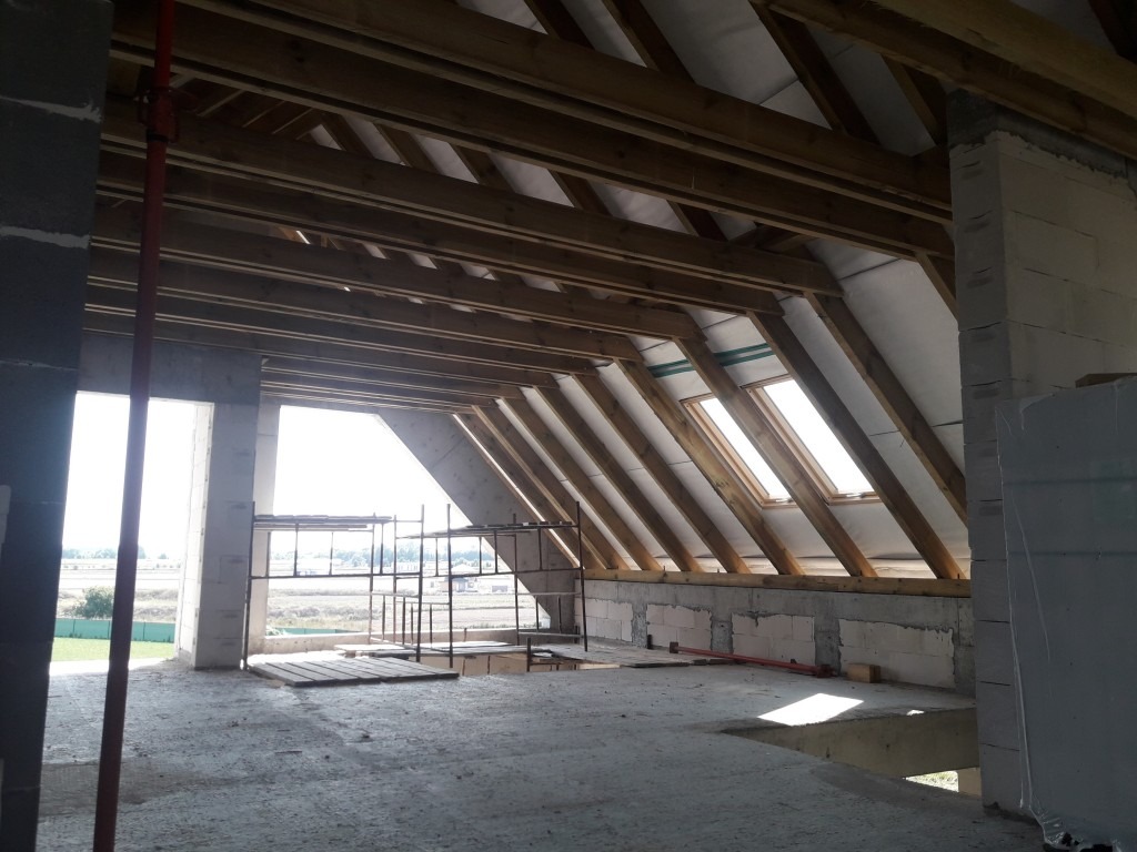 Wnętrze poddasza w trakcie budowy z widoczną konstrukcją drewnianą dachu, oknami połaciowymi i częściowo wymurowanymi ścianami z betonu komórkowego. Na podłodze widoczne rusztowania i deski.