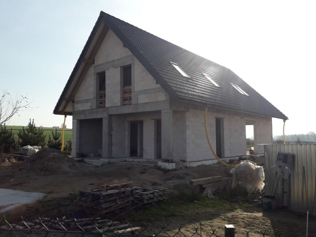 Surowy stan domu jednorodzinnego z białego bloczka, z czarną dachówką i żółtymi rurami wentylacyjnymi, widok z zewnątrz na plac budowy.