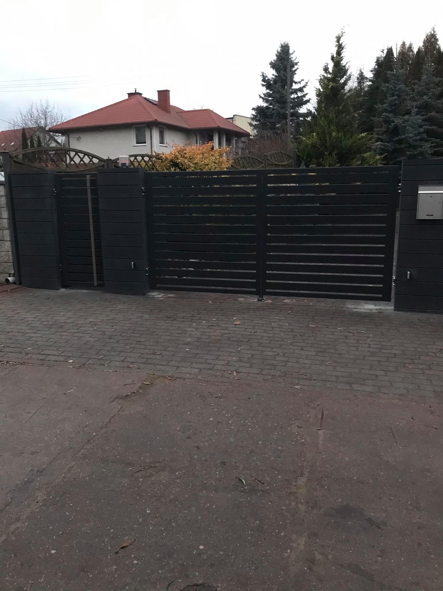 Nowoczesna, pozioma brama wjazdowa z ciemnego drewna, z widocznym fragmentem domu z czerwoną dachówką w tle, na tle zielonych drzew.