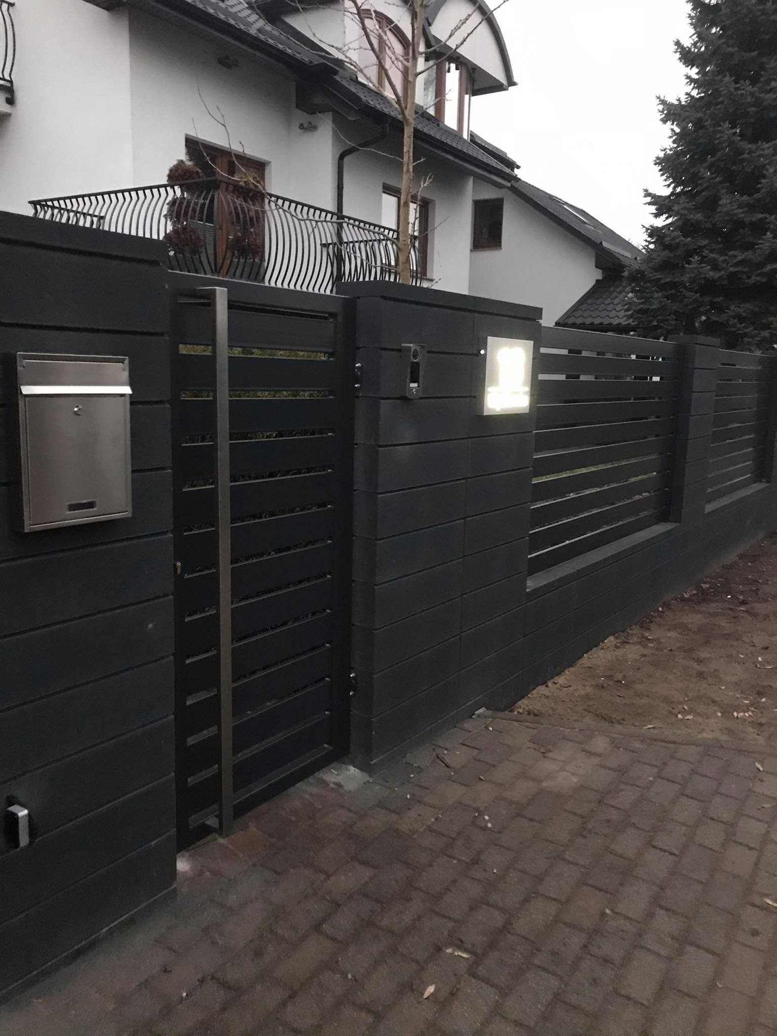 Nowoczesne, grafitowe ogrodzenie posesji z poziomych desek, oświetlony numer domu, skrzynka na listy ze stali nierdzewnej, kostka brukowa na podjeździe, w tle dom z balkonem.