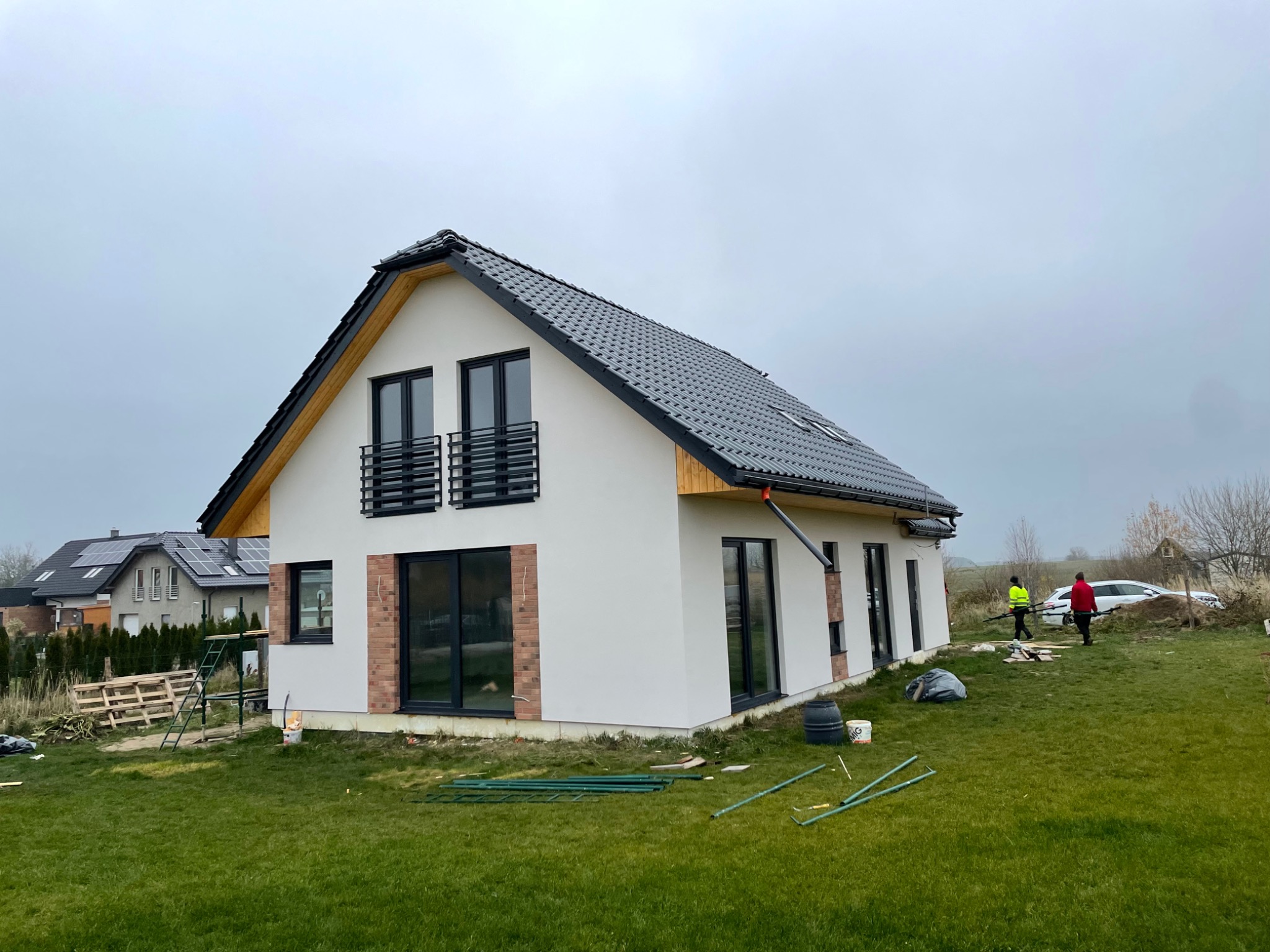 Nowoczesny dom jednorodzinny z dwuspadowym dachem krytym grafitową dachówką, z czarnymi oknami i balkonami, w trakcie wykańczania terenu wokół, na trawniku widoczne narzędzia i materiały budowlane,...