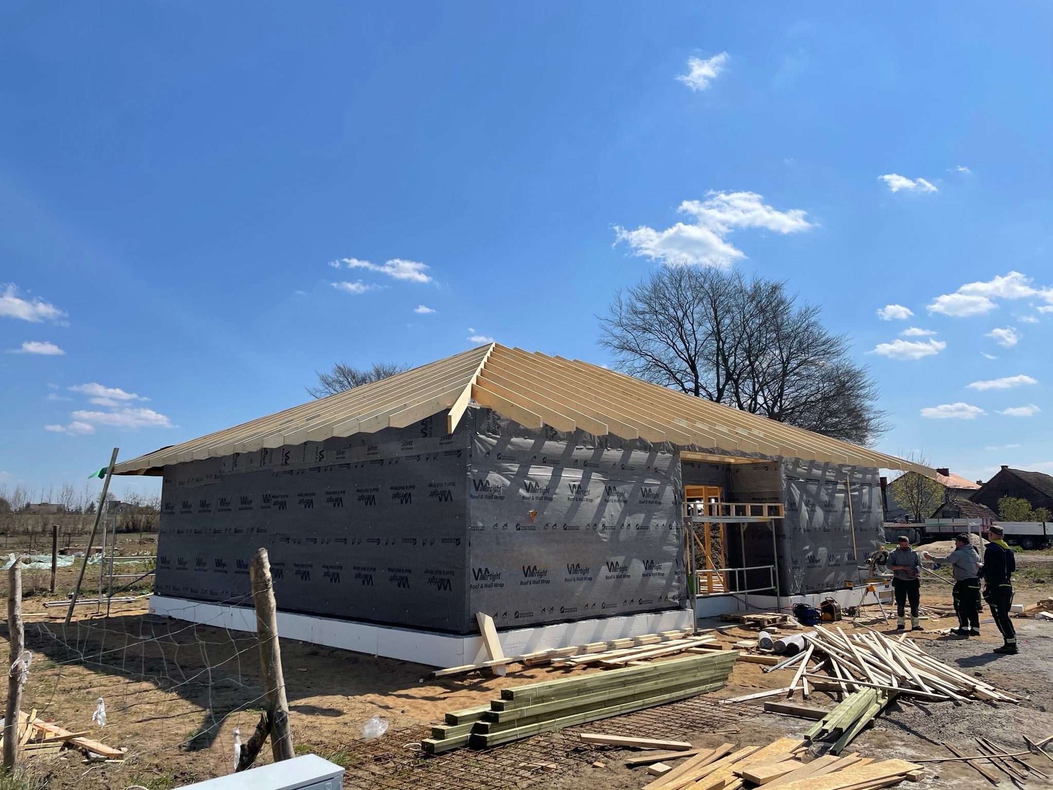 Budowa domu w trakcie wznoszenia konstrukcji dachu; widoczne drewniane elementy więźby dachowej, czarna membrana na ścianach, zgromadzone materiały budowlane i grupa pracowników w oddali.