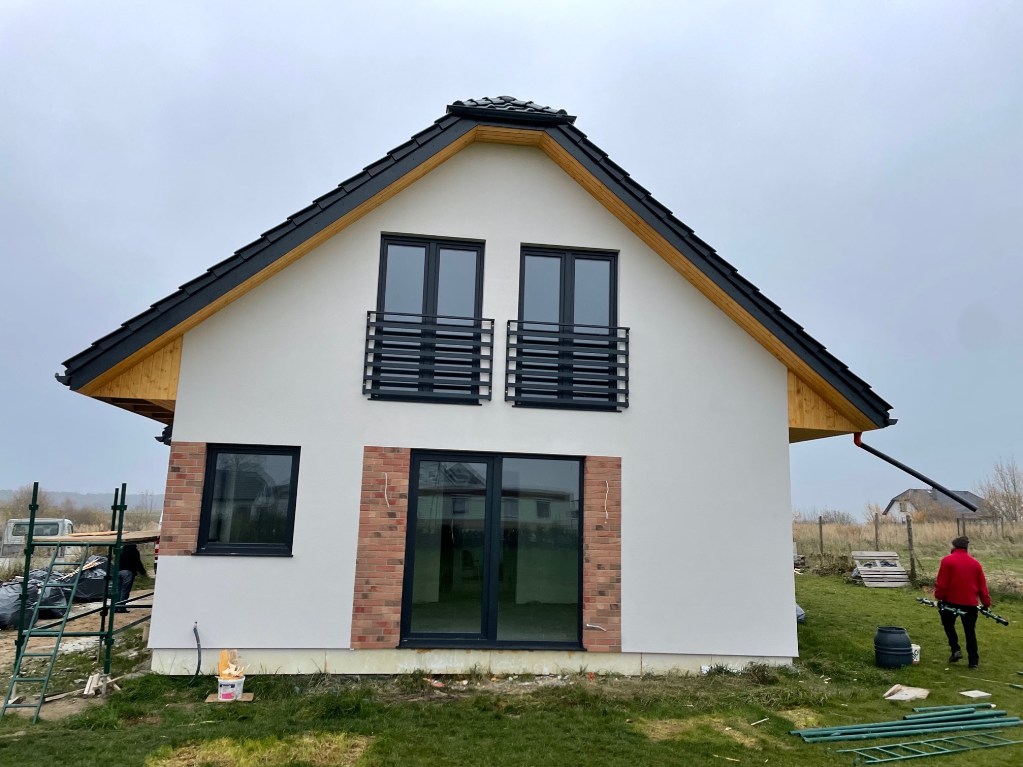 Nowoczesny dom jednorodzinny z białymi ścianami, czarnym dachem i oknami, z fragmentami elewacji wykończonymi czerwoną cegłą. Na pierwszym planie widoczne rusztowania i akcesoria malarskie...