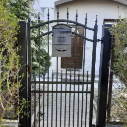 Czarna metalowa furtka z dekoracyjnymi grotami i skrzynką na listy, widok na ścieżkę z kostki brukowej i drzwi wejściowe do domu.