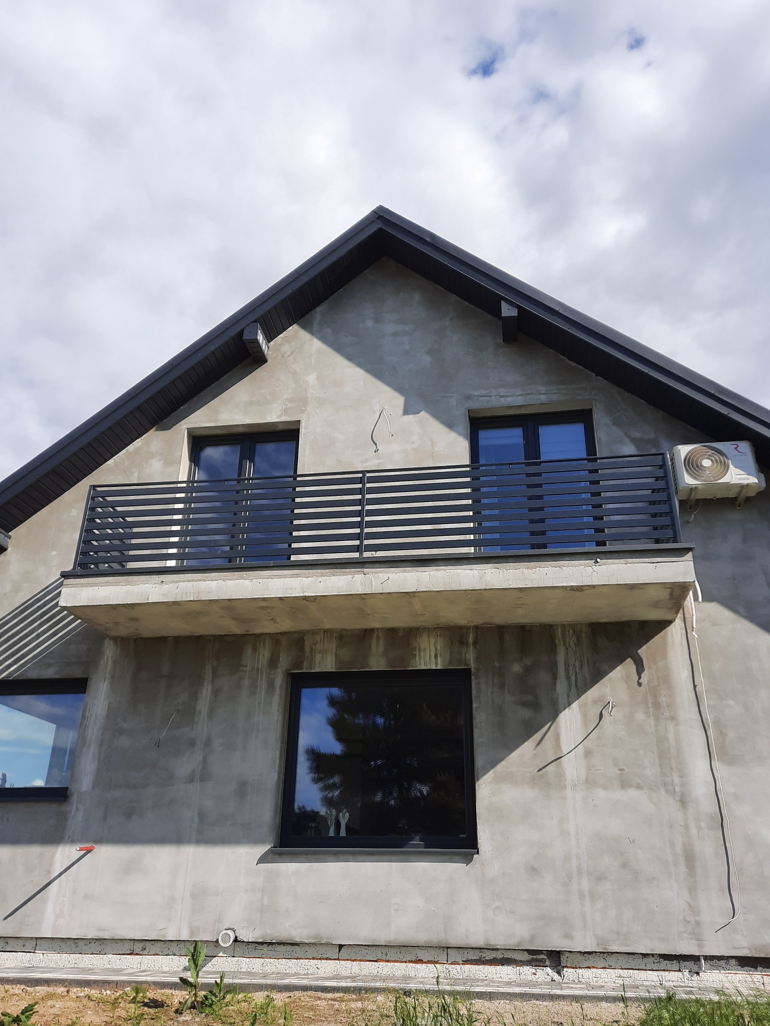 Surowy stan elewacji domu z balkonem zabezpieczonym czarną, poziomą balustradą metalową, widoczny klimatyzator na ścianie balkonu, okna z czarnymi ramami.