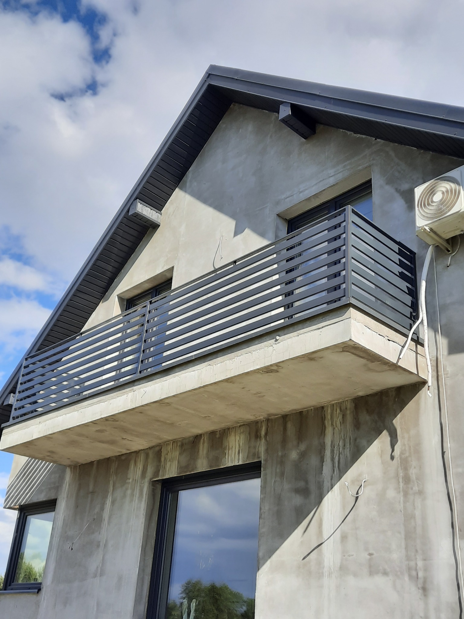 Szary balkon z poziomymi metalowymi balustradami przymocowany do surowej, betonowej ściany budynku z widocznym oknem i jednostką klimatyzacyjną na zewnątrz.