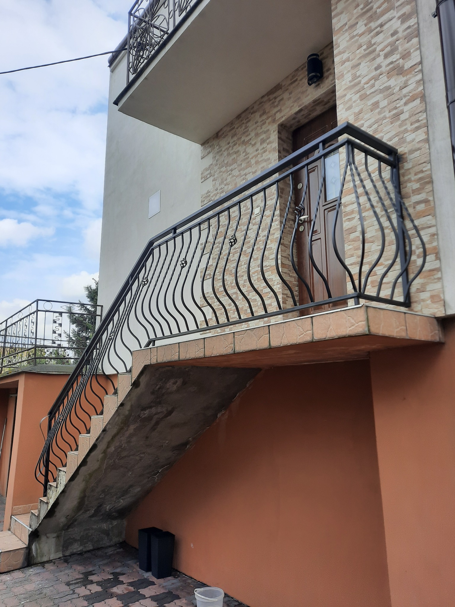 Zewnętrzne schody z kutą, czarną balustradą z motywem fal, prowadzące do drzwi wejściowych z przeszklonym panelem, wykończone płytkami imitującymi kamień, z widocznym fragmentem balkonu z podobną...