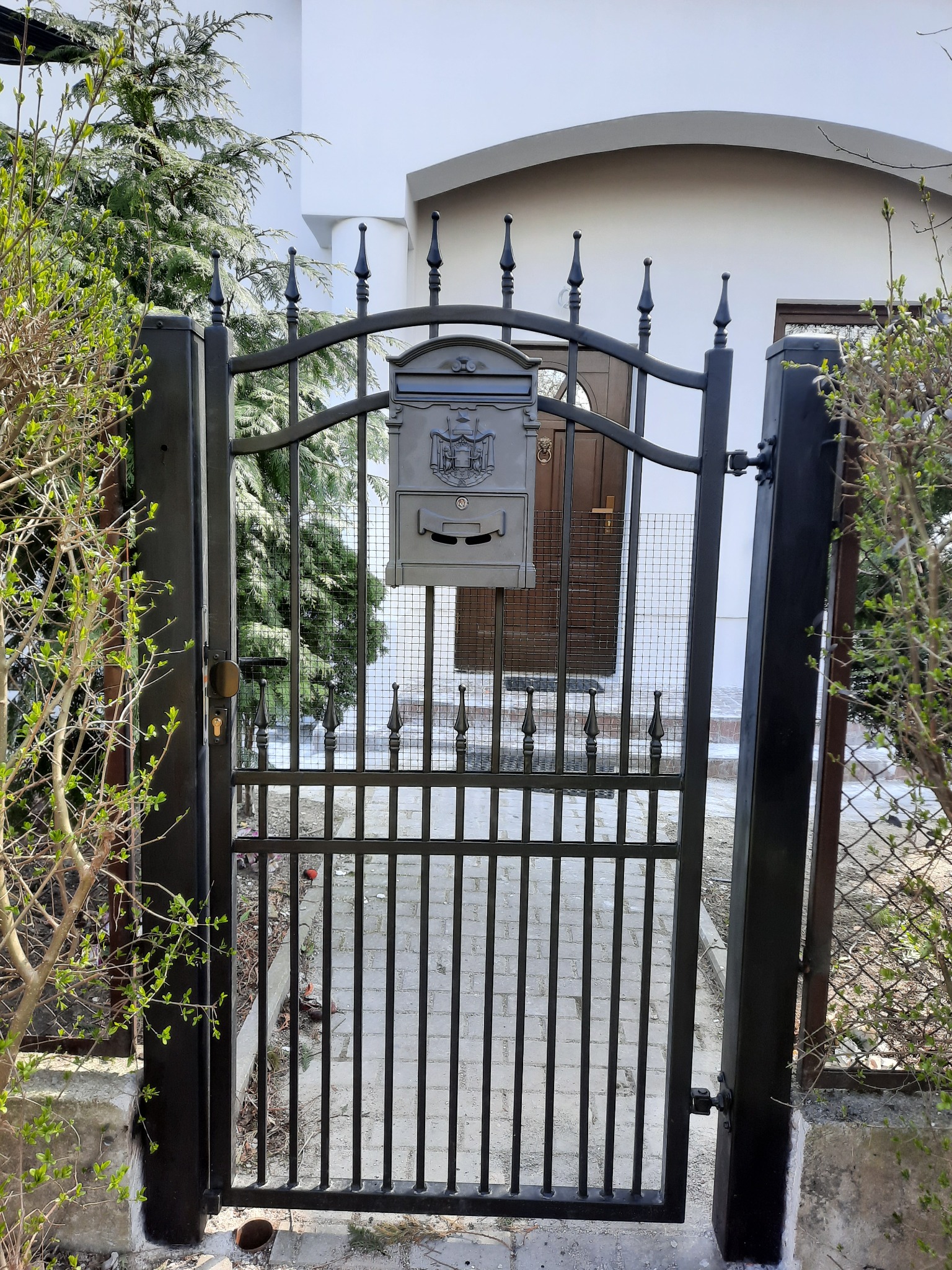 Czarna metalowa furtka z dekoracyjnymi grotami i skrzynką na listy, widok na ścieżkę z kostki brukowej i drzwi wejściowe do domu.