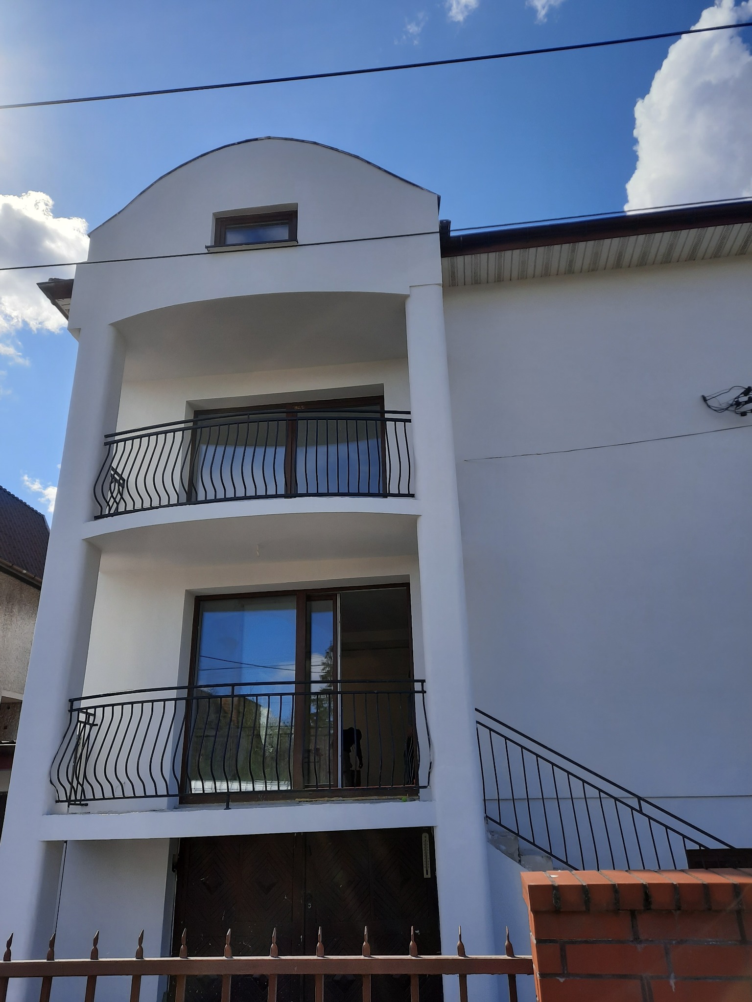 Dwupiętrowy budynek z balkonami z czarnymi, kutymi balustradami, widoczne ogrodzenie posesji i fragment schodów zewnętrznych, błękitne niebo z chmurami w tle.