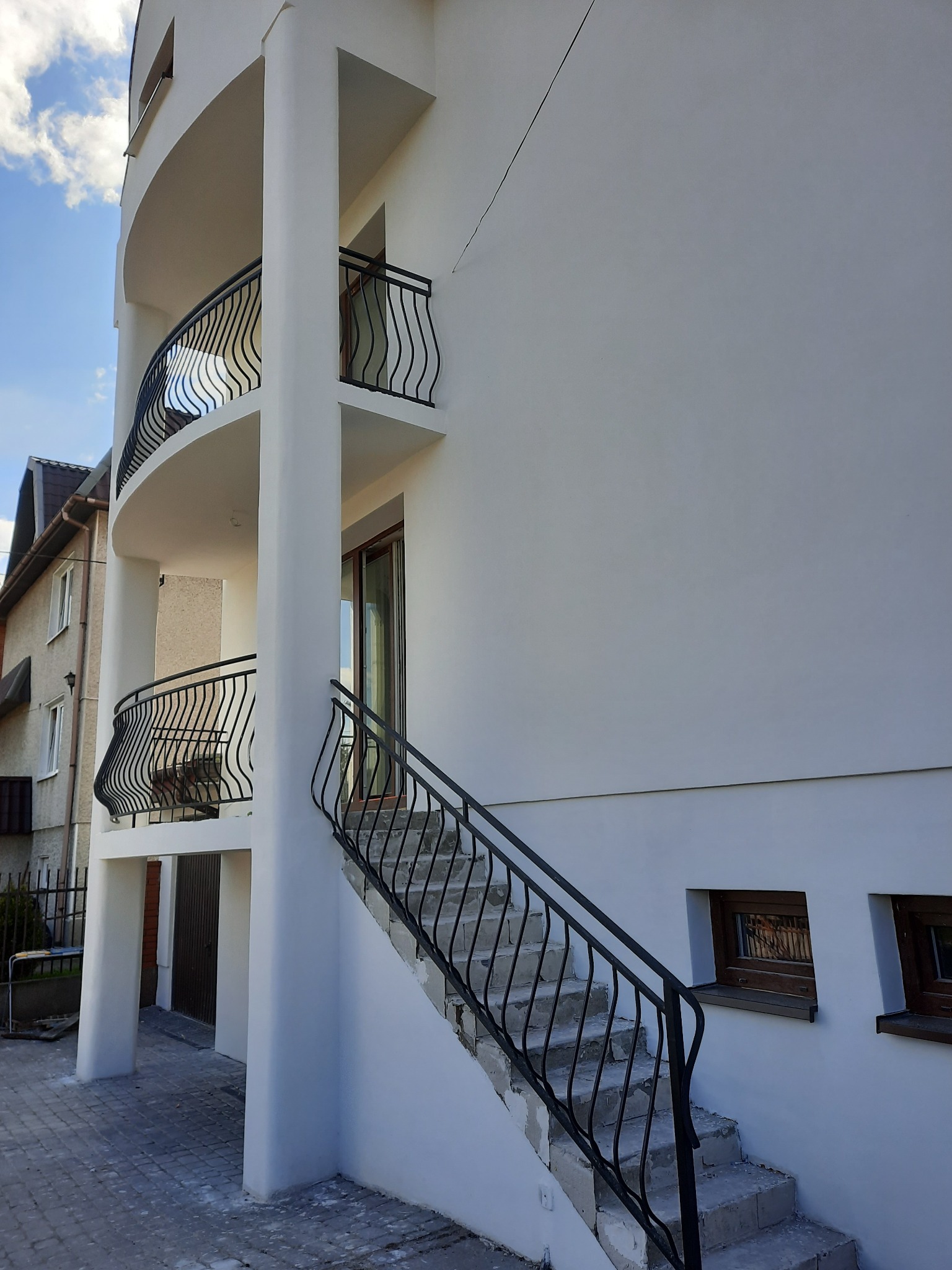 Zewnętrzne schody z czarną, kutą balustradą prowadzące na zaokrąglony balkon nowoczesnego, białego budynku.