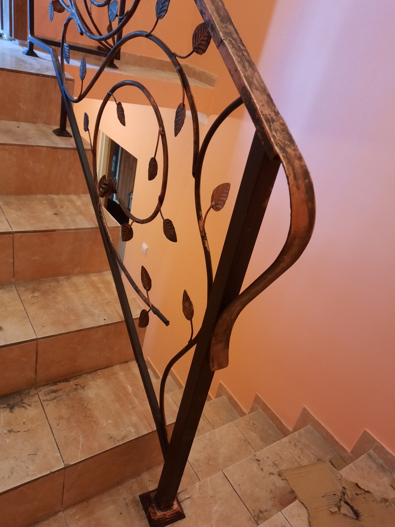 Kute, metalowe balustrady schodowe z motywem liści na tle pomarańczowej ściany i ceramicznych schodów.