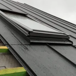 Ciemny metalowy dach z oknem dachowym, widoczne krople wody i fragment konstrukcji drewnianej z zieloną impregnacją.