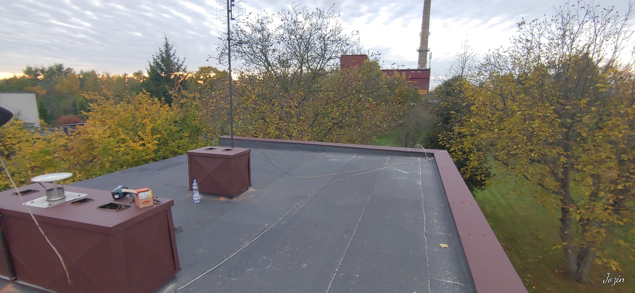 Widok z góry na płaski dach pokryty papą, z dwoma brązowymi kominami wentylacyjnymi, narzędziami i butelką wody, w tle jesienne drzewa i komin przemysłowy.