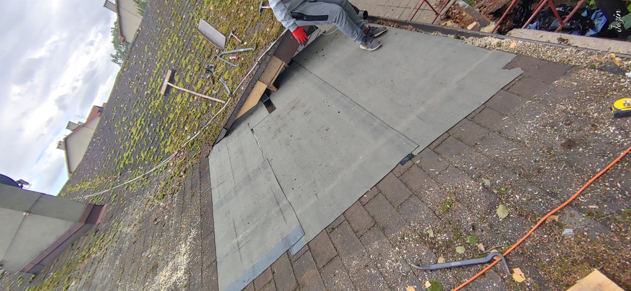 Fragment dachu z papą, częściowo pokryty mchem, narzędzia dekarskie w tle, widoczne nogi dekarza w szarym kombinezonie i czerwonych rękawicach.