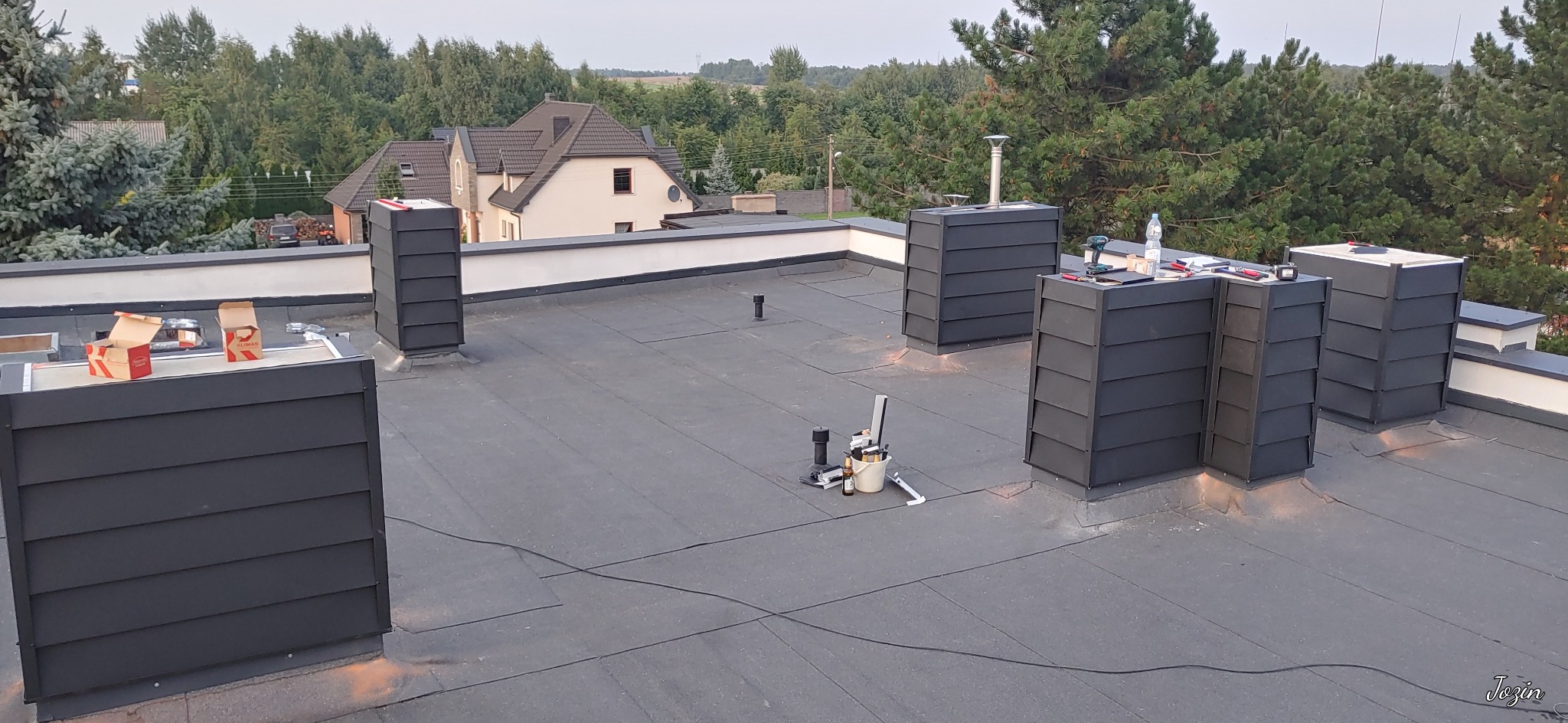 Czarny dach pokryty papą z nowymi obróbkami kominowymi z blachy w kolorze antracytowym, narzędzia pracy i puszki po impregnacie na dachu, w tle dom jednorodzinny i drzewa.