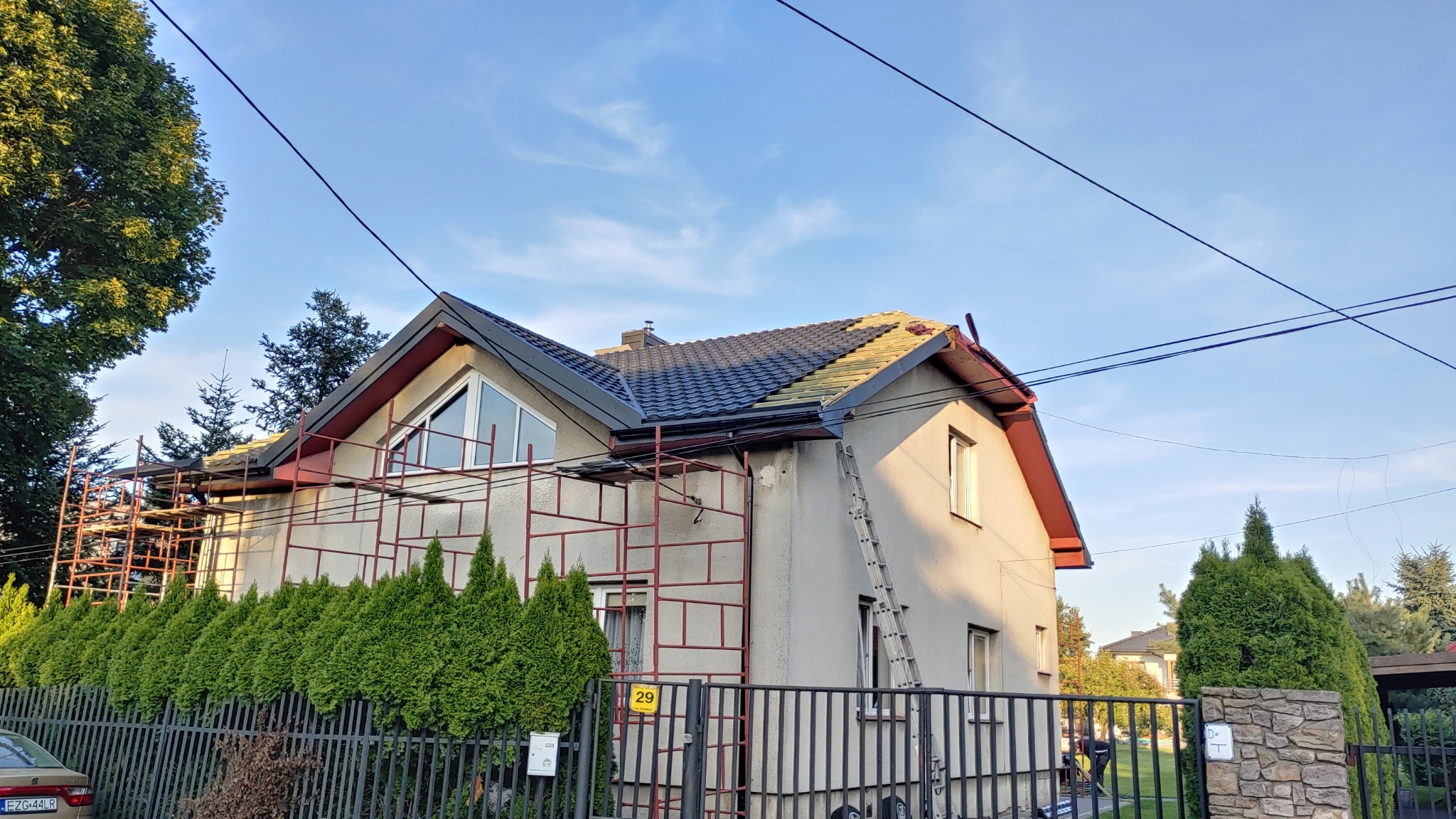 Dom jednorodzinny w trakcie prac dekarskich, widoczne rusztowanie, częściowo położona nowa dachówka w kolorze grafitowym oraz stara, jasna dachówka, drabina oparta o ścianę budynku, błękitne niebo...