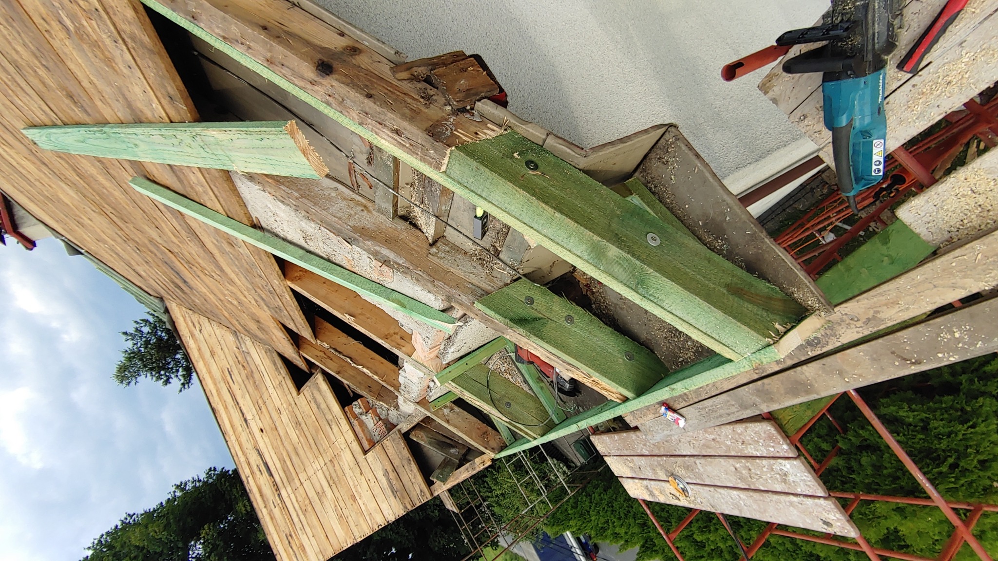 Wymiana elementów więźby dachowej z użyciem zielonych, impregnowanych belek, widoczna piła łańcuchowa i fragment nieba.