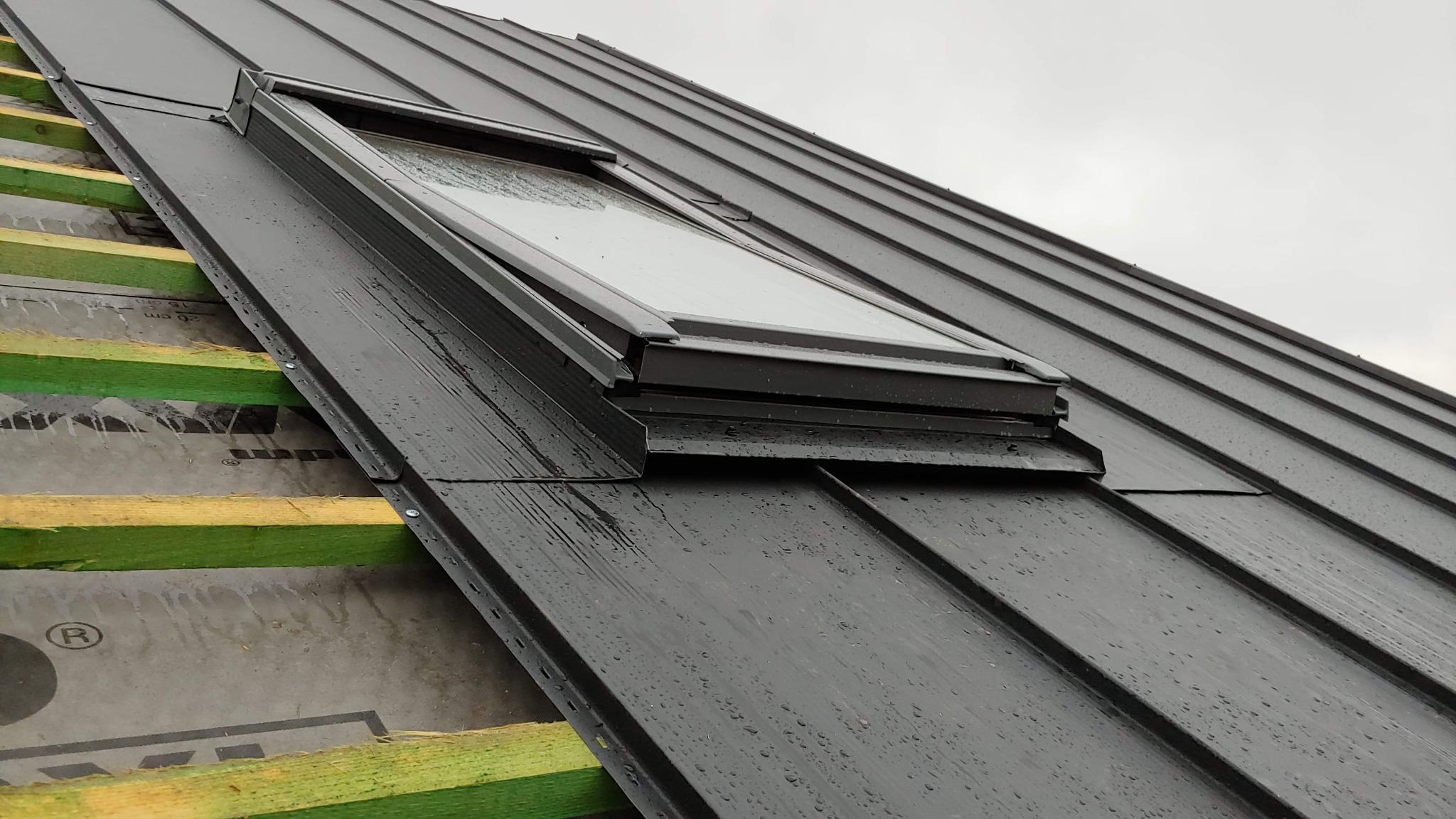 Ciemny metalowy dach z oknem dachowym, widoczne krople wody i fragment konstrukcji drewnianej z zieloną impregnacją.