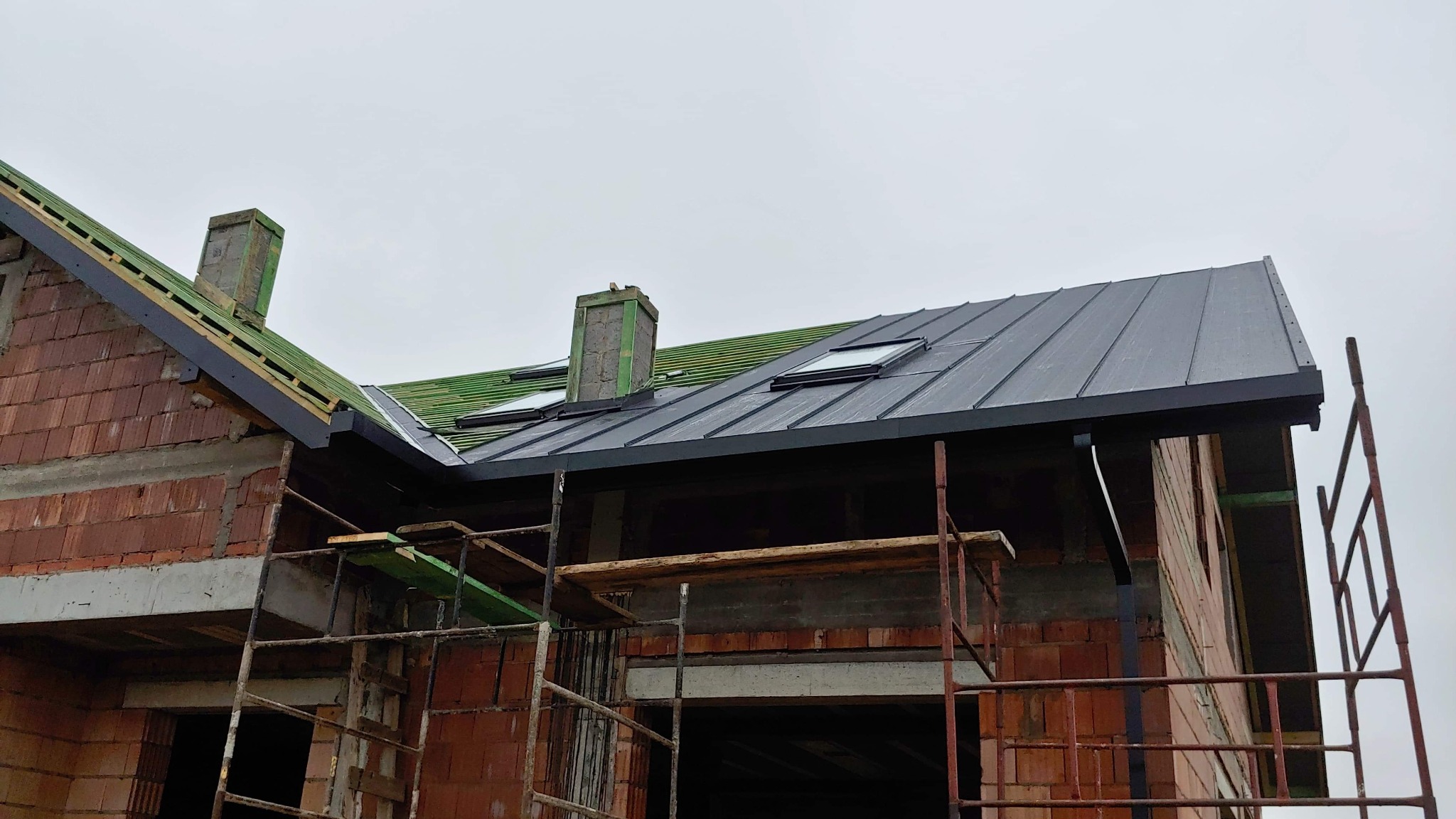 Budowa domu z nowym, grafitowym dachem z oknami połaciowymi, widoczne rusztowania i komin w trakcie wykańczania.